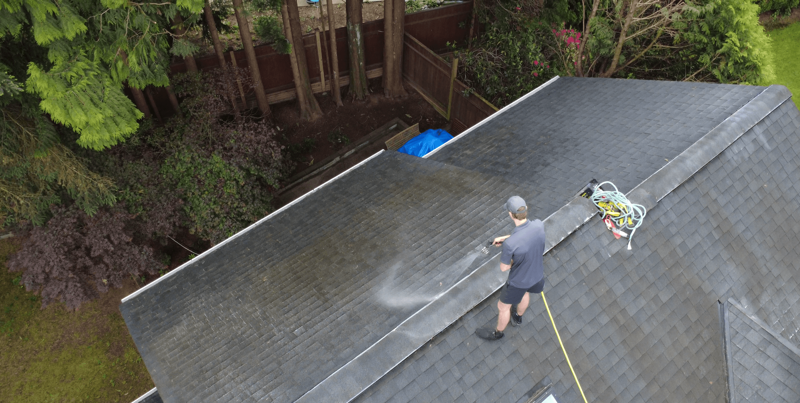 Aerial view of a worker cleaning a dark shingle roof with a pressure washer.